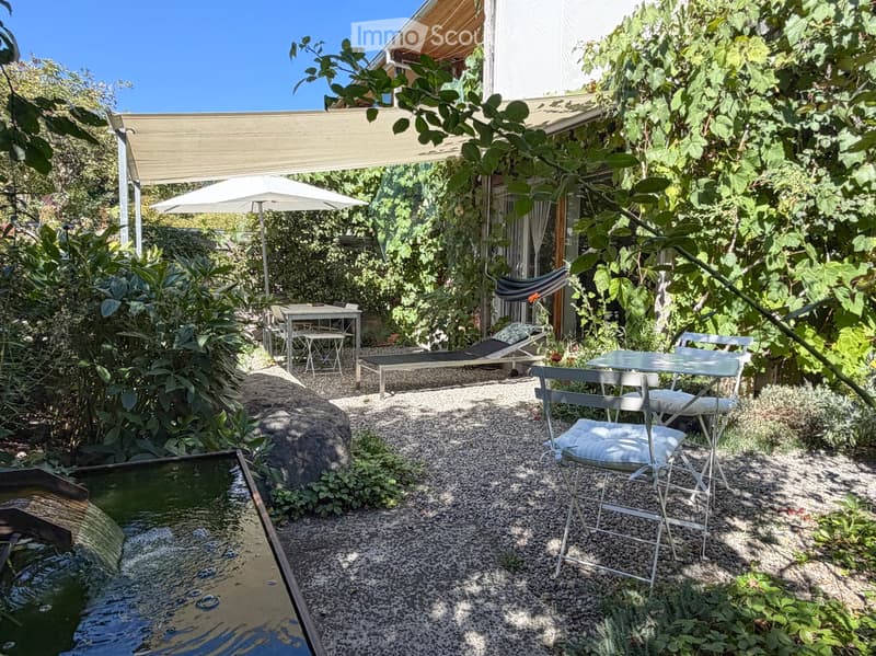 A lush garden patio with a small water feature, lounge chairs, tables, and umbrellas, surrounded by greenery and a covered seating area.