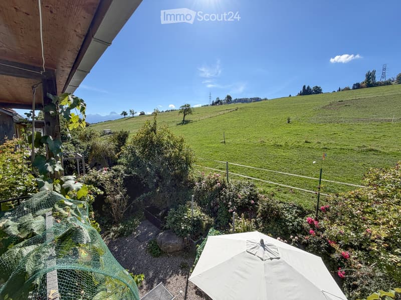 Garden with a white umbrella, plants, and a scenic view of green hills and mountains under a clear blue sky.