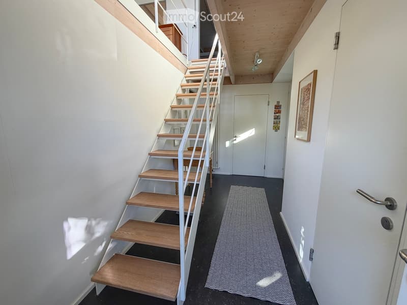 A modern hallway with a wooden staircase, white walls, and a grey rug on the floor. A framed picture hangs on the wall, and a door is visible at the end of the hallway.