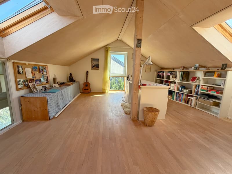 Bright attic room with wooden floors, slanted ceilings, large windows, and skylights. Features a bookshelf, a small table with a cushion, a guitar, and a wicker basket.