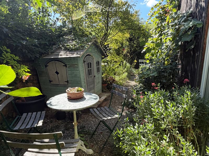 A small, lush garden with a round table, chairs, a flower pot, and a small wooden toy house surrounded by greenery and plants.