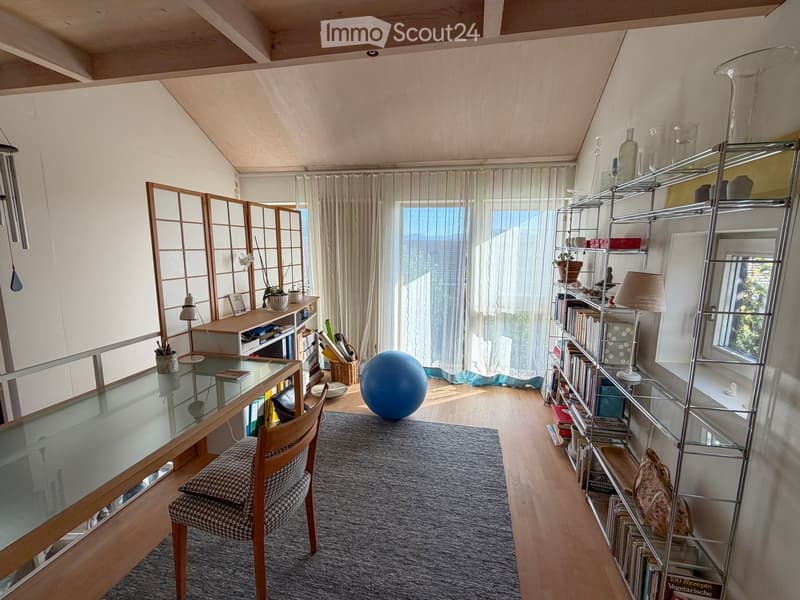 A modern, well-lit room with wooden floors, large windows, a glass table, a chair, a shelf with books and decorative items, and a blue exercise ball.