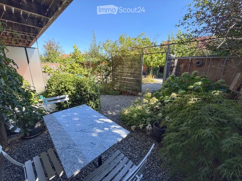 A small, well-maintained garden with a stone table and chairs, surrounded by lush greenery and a wooden fence.