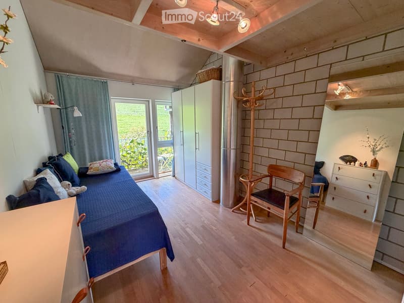 Cozy bedroom with wooden flooring, a blue bed, a wooden chair, a wardrobe, and a coat rack. A balcony is visible through the open door.