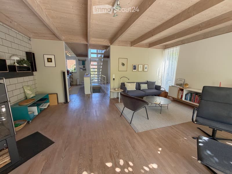 Modern living room with wooden floors, exposed wooden ceiling beams, a grey sofa, coffee table, armchairs, bookshelf, and fireplace.