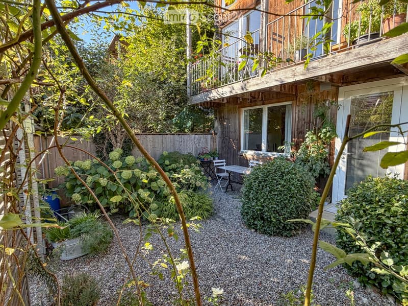 A small, well-maintained garden area with gravel ground, various plants, shrubs, and a small seating area with a table and chairs near the building entrance.