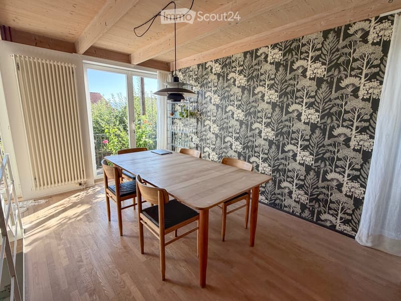 Dining room with wooden floor, dark patterned wallpaper, large window with view, dining table with chairs, pendant light, radiator, and shelf with plants.