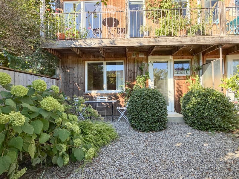 Garden area with gravel ground, small table and chairs, lush greenery, and a wooden house with a balcony above.