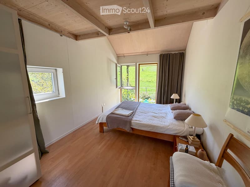 Cozy bedroom with wooden floors, a double bed with pillows and blankets, a nightstand with a lamp, a small wooden chair, and large windows with curtains offering a view of the outdoors.