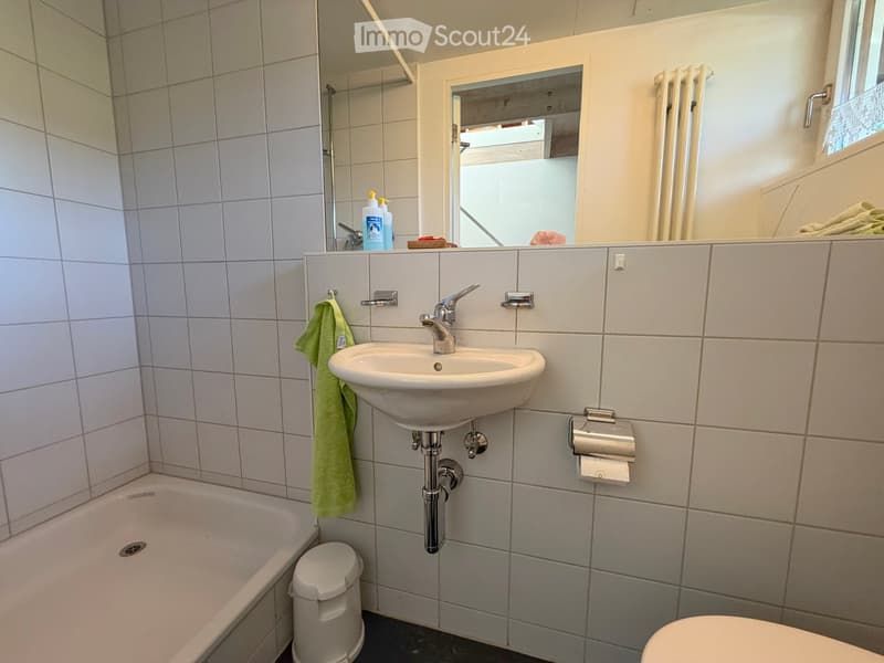 Modern bathroom with white tiled walls, a sink with a chrome faucet, a bathtub, a toilet, a towel, and a soap dispenser on the counter.