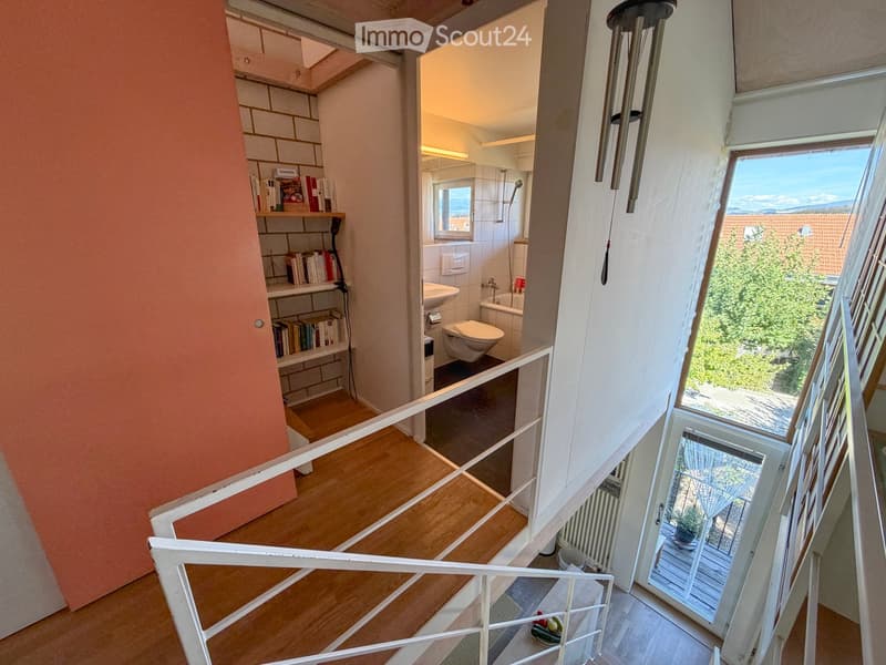 Spacious hallway with wooden flooring, white railing, open staircase leading to a bathroom with white tiles, sink, toilet, and bathtub. A window offers a scenic view of greenery and rooftops.