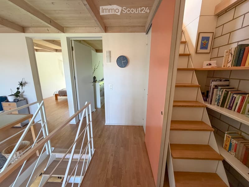 A modern hallway with wooden flooring, white walls, and a staircase leading to an upper level. The hallway features a clock on the wall, a small shelf with books, and a view into an adjacent living area with a bed and a small table.