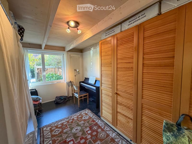 A cozy, well-lit room with wooden ceilings and walls, featuring a piano, a chair, a curtain, a radiator, a lamp, and a patterned rug.