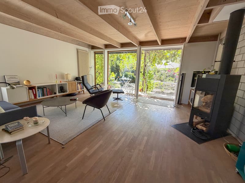 Modern living room with wooden flooring, exposed wooden ceiling beams, large glass doors opening to a garden, fireplace, sofa, coffee table, chairs, bookshelf, and decorative items.