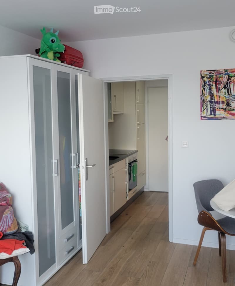 A furnished room with a wooden floor, a white wardrobe with glass doors, a small table with a chair, and a colorful painting on the wall. A small kitchen area is visible through an open doorway.