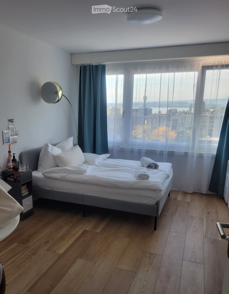 Cozy bedroom with a large bed, nightstand, floor lamp, and a scenic view through large windows.