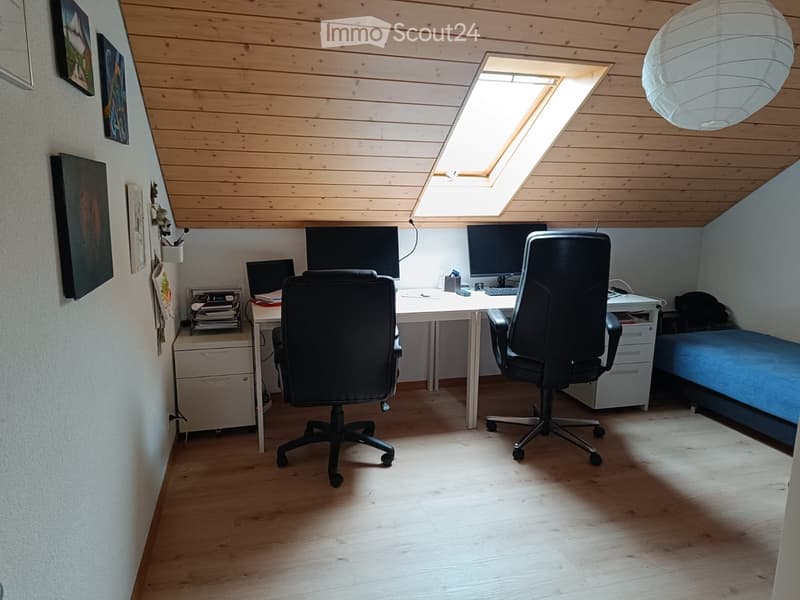 A multi-functional room with a desk setup and a bed, featuring wooden flooring, a skylight, and a hanging lantern.