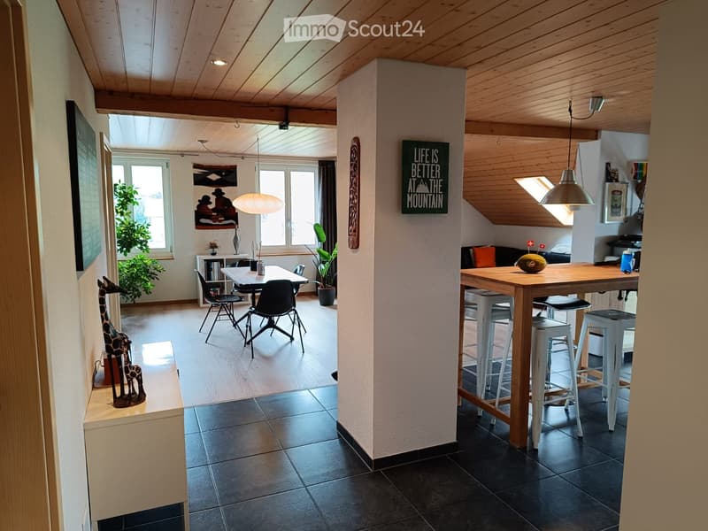 Open-plan living and dining area with wooden beams, pendant lights, and large windows. Includes a dining table with chairs, a kitchen island with stools, a sofa, and decorative items.