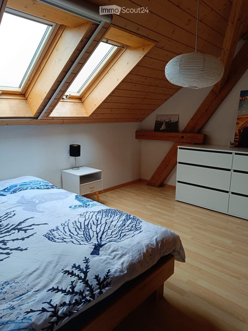 Cozy attic bedroom with wooden slanted ceilings, skylights, a bed with blue and white patterned bedding, a white nightstand with a lamp, a white dresser, and a hanging pendant light.