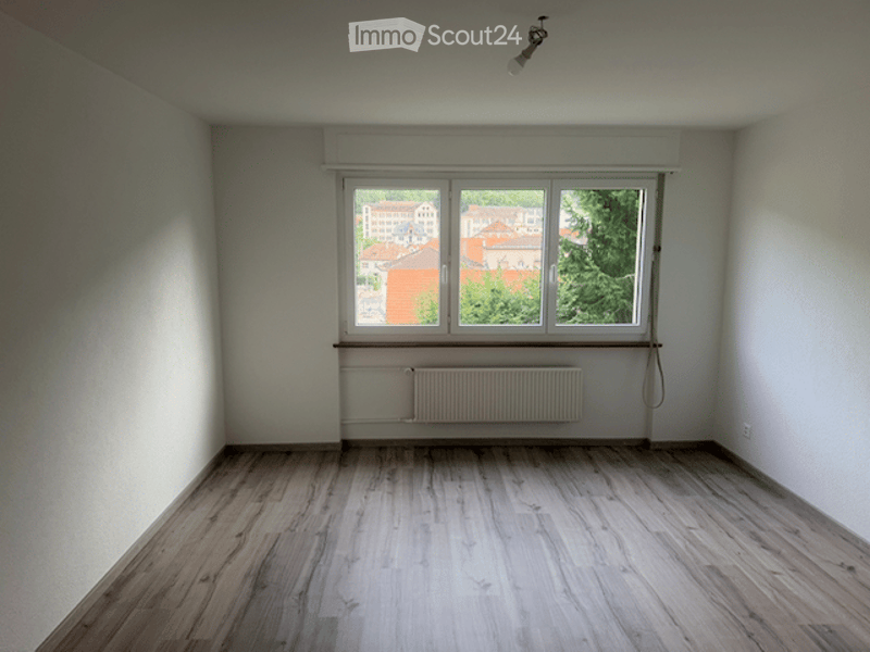 Empty room with wooden flooring, white walls, large window with view, radiator, and ceiling light fixture.
