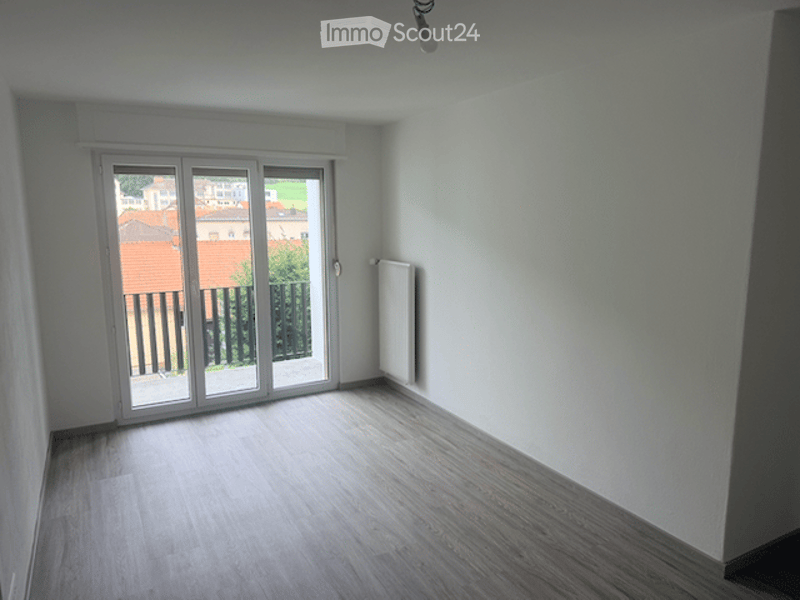 Empty room with wooden flooring, white walls, large sliding glass door leading to a balcony, radiator on the wall, and a view of the outdoor area.