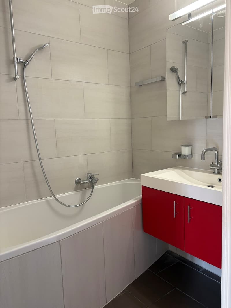 Modern bathroom with a white bathtub, red cabinet, white sink, mirror, and showerhead.