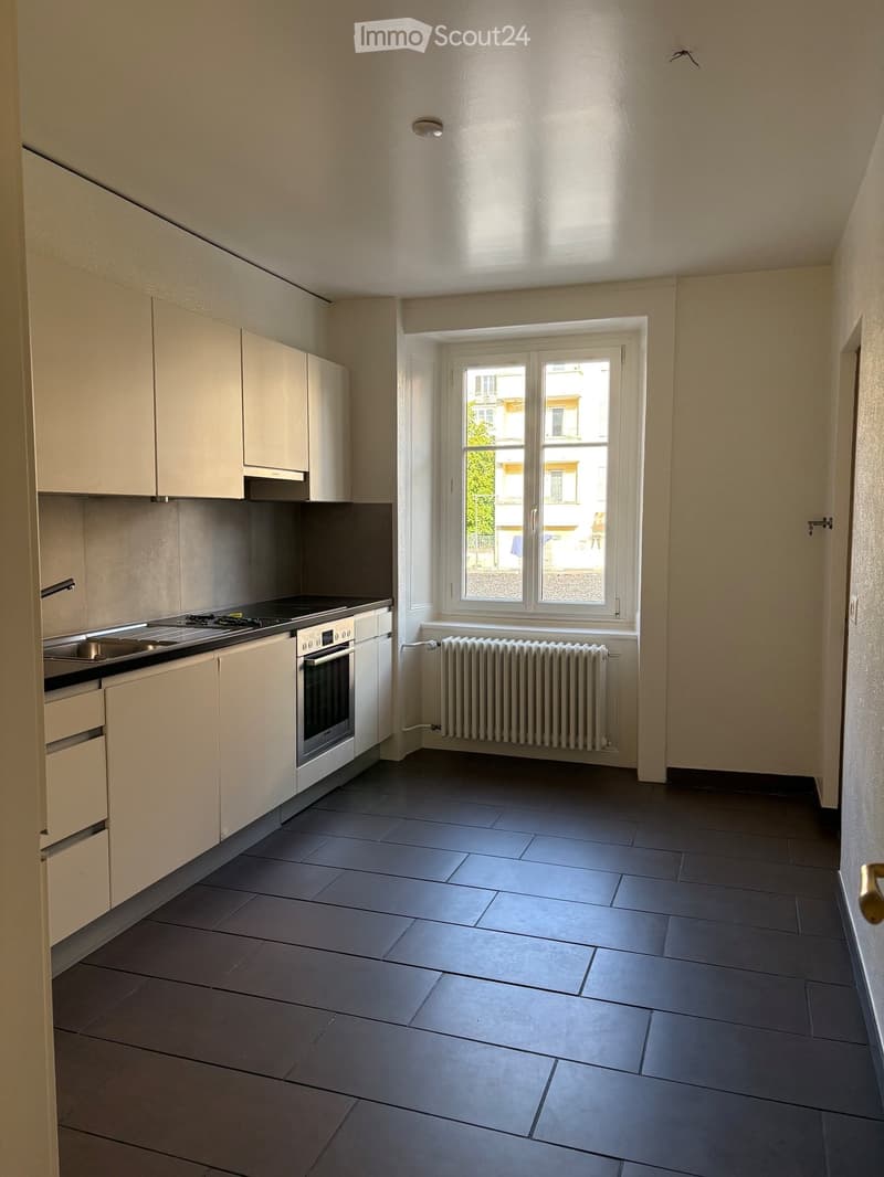 Modern kitchen with light-colored cabinets, a stove, oven, sink, and radiator under a large window.