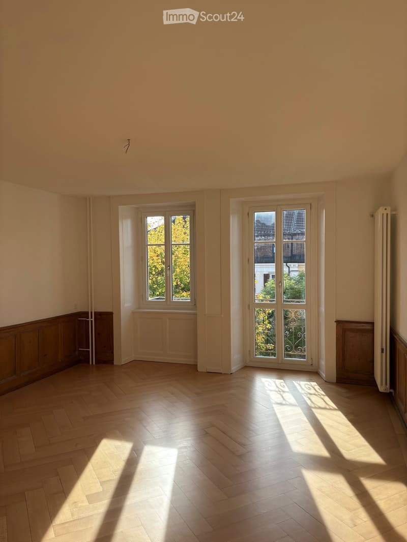 Empty room with light wooden flooring, large windows, and cream-colored walls. Natural light streams in, casting shadows on the floor.