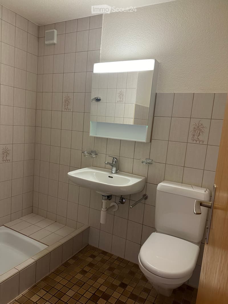 Bathroom with beige tiled walls, a white sink with a chrome faucet, a toilet, a bathtub, and a mirror with a built-in light. The floor has a patterned tile design.