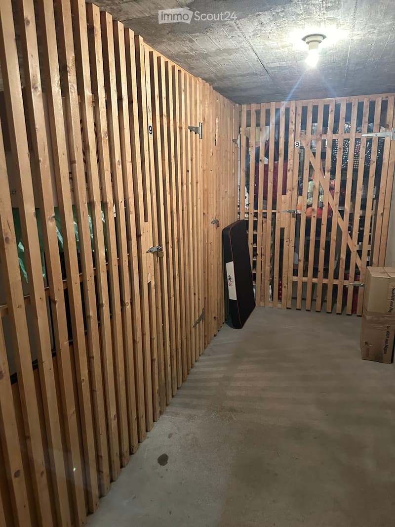 A narrow, unfinished hallway with wooden paneling on both sides, concrete ceiling, and a concrete floor. The hallway features numbered doors and a storage area with boxes and a folded garment bag visible at the end.