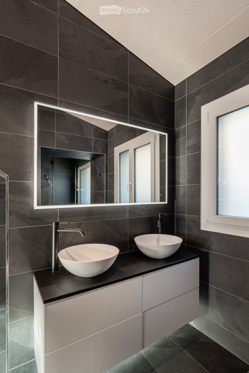 Modern bathroom with dark tiled walls and floor, two white bowl sinks, sleek faucets, and a large illuminated mirror.