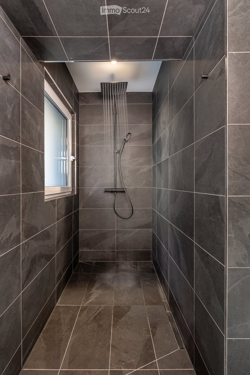 Modern shower area with dark grey tiles, a large overhead showerhead, and a handheld shower hose. The space features a sleek, contemporary design with a window on one side.