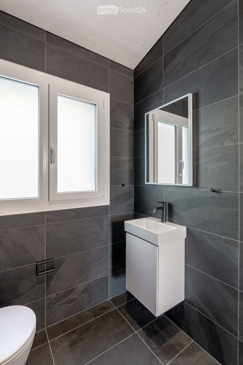 Modern bathroom with dark grey tiles, white sink, mirror, and toilet.