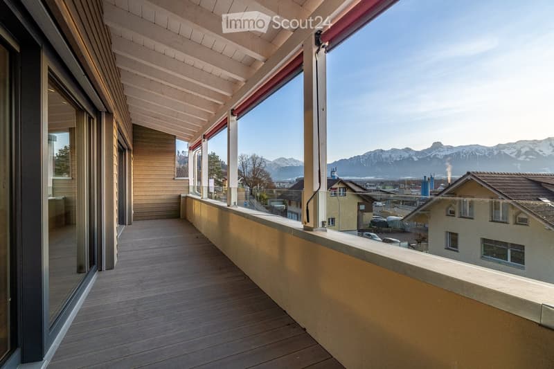 Balcony with wooden decking, covered by a wooden pergola, overlooking a town with distant snow-capped mountains.