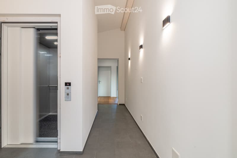 Modern hallway with white walls, dark tiled floor, and recessed lighting. Features an elevator on the left and a door leading to another room on the right.