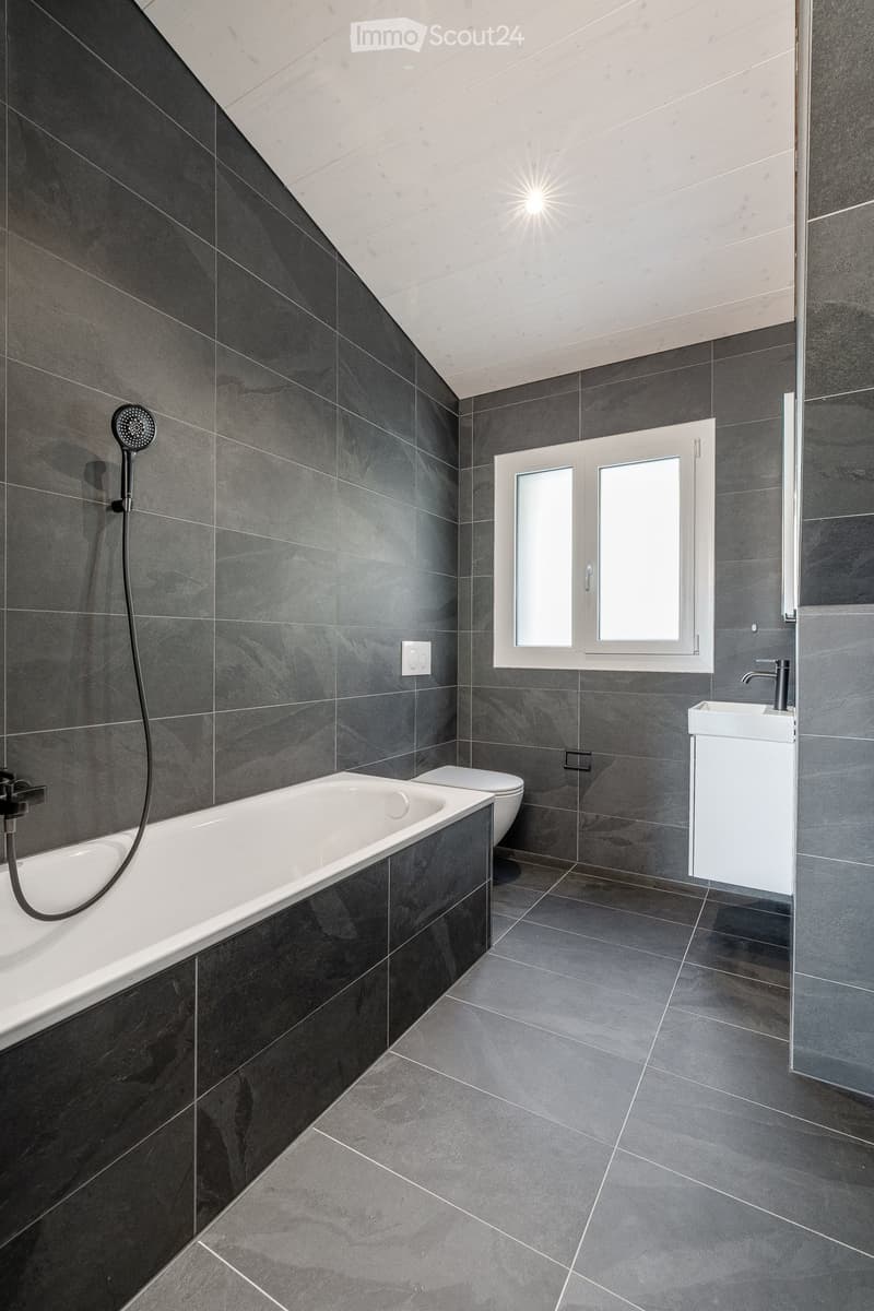 Modern bathroom with dark grey tiles, a white bathtub, a toilet, a white sink with a chrome faucet, and a showerhead.