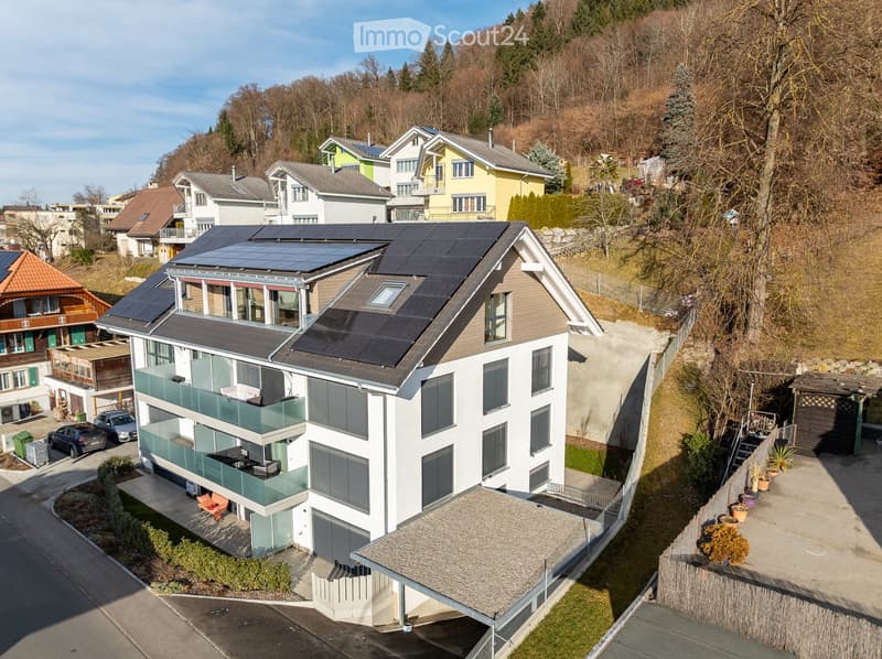 Modern two-story house with solar panels on the roof, large windows, balconies, and a garage. Surrounded by other houses and greenery.