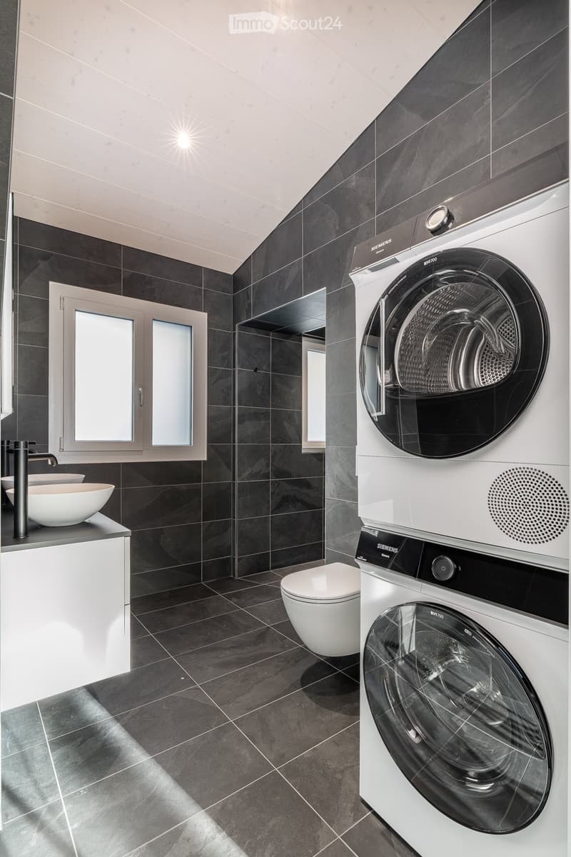 Modern bathroom with dark tiled walls and floor, white sink, faucet, toilet, and stacked Siemens washing machine and dryer.