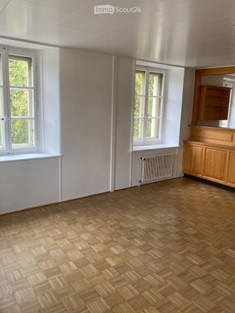 Empty room with wooden herringbone flooring, white walls, two windows, radiator, and built-in wooden cabinetry.