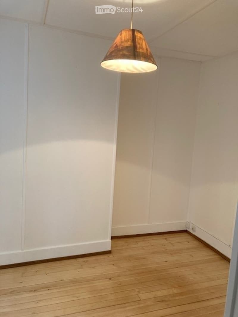Completely empty room with white walls and wooden floor. A single hanging lamp is visible on the ceiling.