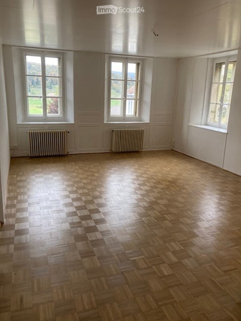 Empty room with wooden parquet flooring, white walls, and three large windows offering a view of a green landscape. Radiators are visible under the windows.