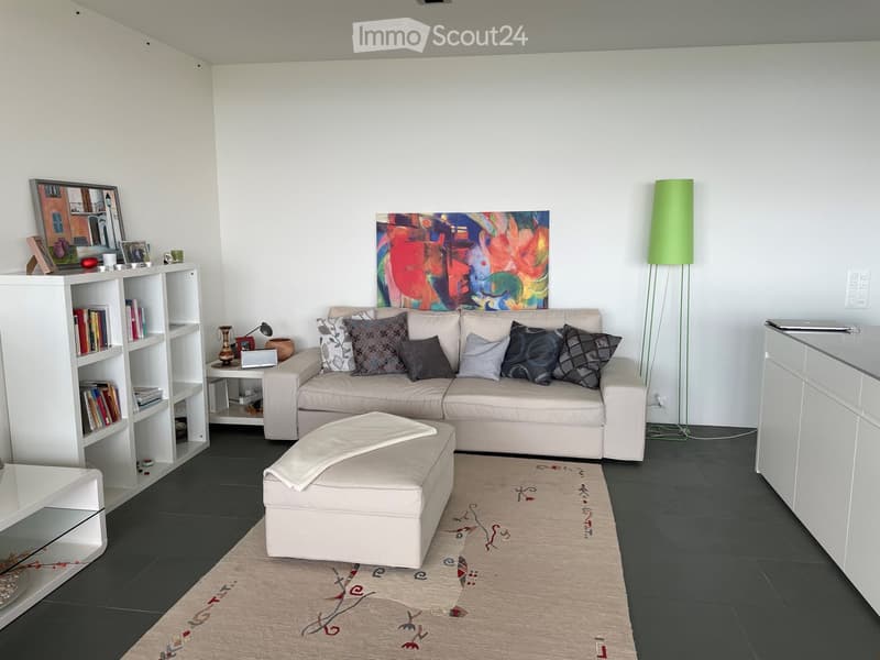 Modern living room with a light-colored sectional sofa, ottoman, bookshelf, small table, floor lamp, and artwork on the wall.