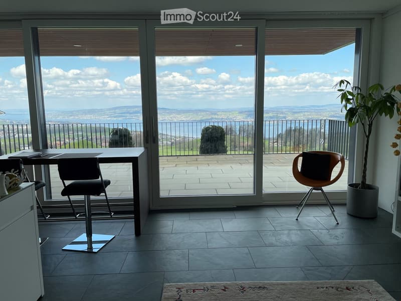 Bright room with large sliding glass doors opening to a balcony, offering scenic views. Includes a table, chairs, a stool, and a potted plant.