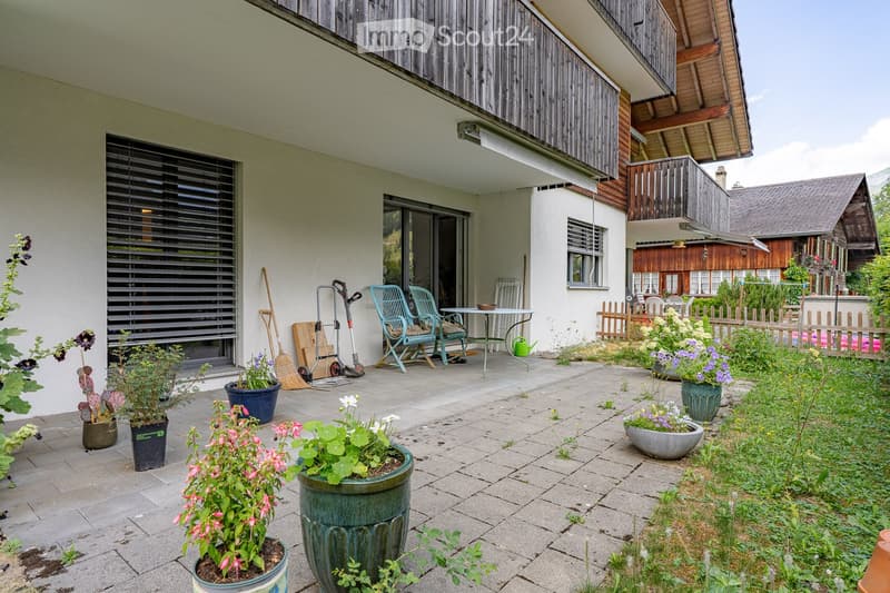 A well-maintained garden area with potted plants, a small table and chairs, a broom, and a garden hose. The space is paved with stone tiles and features a wooden fence in the background.