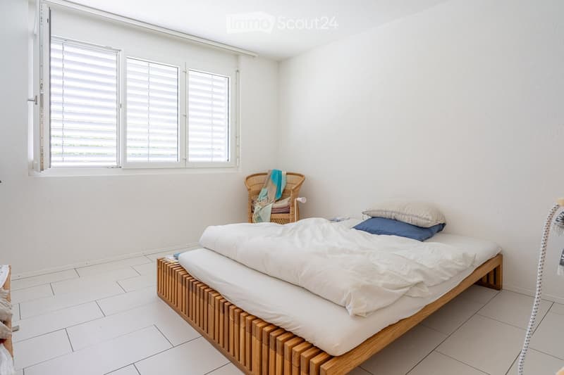 A modern, minimalist bedroom with a wooden bed, white bedding, a wicker chair, and large windows with blinds.