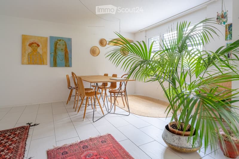 Bright room with tiled floor, a wooden dining table surrounded by chairs, large potted plant, and two paintings on the wall.