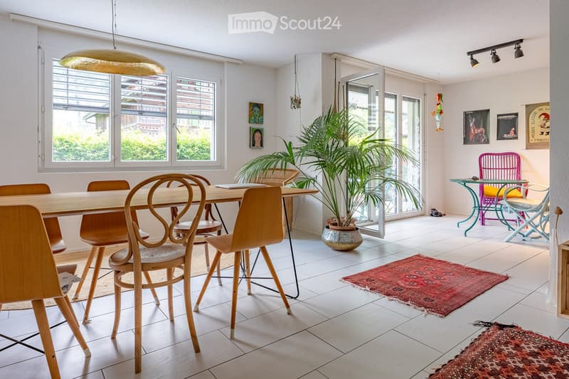 Bright dining area with a wooden dining table and chairs, a potted plant, and colorful side chairs near large windows.