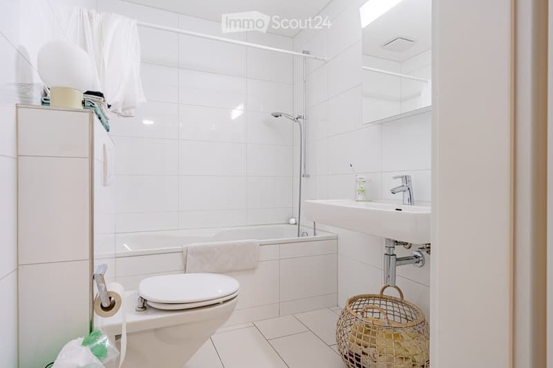Modern bathroom with white tiles, bathtub, toilet, sink, mirror, and a wicker basket on the floor.