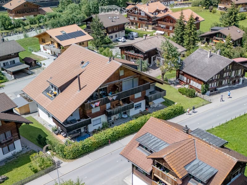 Aerial view of a residential area with multiple houses featuring wooden facades, tiled roofs, and balconies. The houses are surrounded by greenery and have driveways with parked cars.