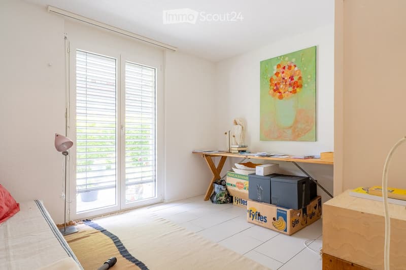 Bright room with white walls, tiled floor, large sliding glass doors leading to a balcony, a wooden table with books and a painting on the wall, and a floor lamp near the entrance.